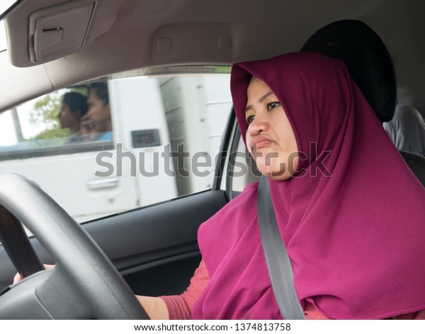 Portrait of funny Asian female muslim driver get\
bored in her car trapped in traffic jam, tired lazy facial\
expression gesture