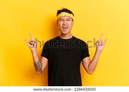 Portrait of funny asian man in black t-shirt and yellow bandana posing on yellow studio background, expression of joy on his face, shows little finger and forefinger put forward sign