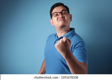 Portrait of funny Asian man showing cynical unhappy angry facial expression putting up his fist challenge to fight, against blue background