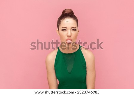 Portrait of funny attractive woman with bun hairstyle, showing tongue out, demonstrating childish behavior, wearing green dress. Indoor studio shot isolated on pink background.