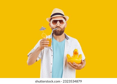 Portrait of a funny bearded man in beach hat and sunglasses drinking orange juice cocktail holding inflatable duck isolated on yellow studio background. Summer holiday trip and vacation concept - Powered by Shutterstock