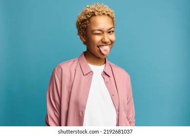 Portrait of funny black girl with blond curly hair showing tongue winking with one eye, making faces, fooling around, aping with silly face, joking, teasing, isolated on blue studio background