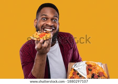 Portrait Of Funny Black Guy Eating Pizza Slice Smiling To Camera Overeating Posing In Studio Over Yellow Background. Male Foodie Enjoying Junk Food. Unhealthy Nutrition And Cheat Meal Concept