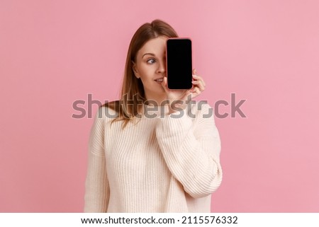 Portrait of funny blond woman covering her eye with mobile phone with empty screen for advertisement, looking away, wearing white sweater. Indoor studio shot isolated on pink background.