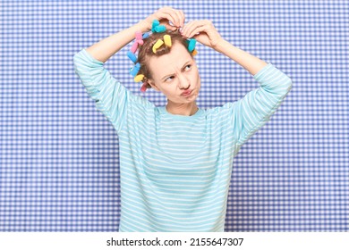 Portrait of funny blond young woman putting her hair into bright colorful curlers, looking busy, frowning face, standing over shower curtain background in bathroom. Beauty and morning routine concept