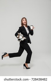 Portrait Of A Funny Busy Businesswoman In Suit Holding Take Away Coffee Cup And A Newspaper While Running Late And Looking At Camera Isolated Over White Background