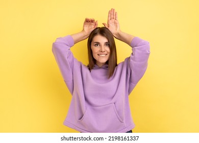 Portrait Of Funny Carefree Attractive Woman Showing Bunny Ears Gesture And Looking Playful Happy, Childish Behavior, Wearing Purple Hoodie. Indoor Studio Shot Isolated On Yellow Background.