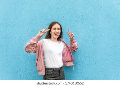 Portrait Of Funny Caucasian Teen Girl In Blank White T-shirt And Pastel Pink Bomber Jacket Against Blue City Wall Solid Background. 