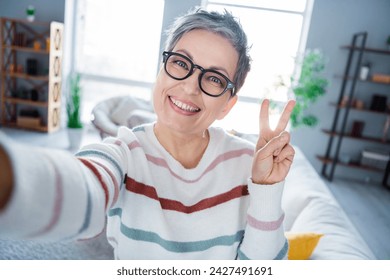 Portrait of funny cheerful person beaming smile make selfie hand fingers demonstrate v-sign modern interior flat indoors