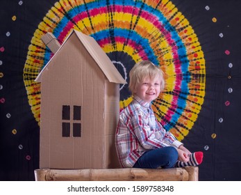 Portrait Of Funny Child In Glasses For Vision And Plaid Shirt.  Boy, With The Help Of His Parents, Built Cardboard House. Kid Is Pleased With Work Sitting Next To Him And Enjoys Toy House