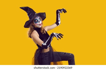 Retrato de niño gracioso en disfraz de Halloween bailando aislado con fondo de color amarillo. Niña feliz con disfraz de esqueleto y sombrero de bruja, con maquillaje de cráneo divirtiéndose en la fiesta de Halloween