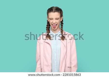 Portrait of funny childish teenager girl with braids wearing pink jacket standing with closed eyes, sticking her tongue out, having fun. Indoor studio shot isolated on green background.