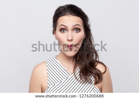 Portrait of funny crazy beautiful young brunette woman with makeup and striped dress standing and looking at camera with big eyes and fish lips gesture. indoor studio shot, isolated on grey background