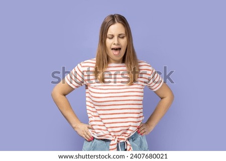 Portrait of funny crazy blond woman wearing striped T-shirt standing with closed eyes and showing tongue out, keeps hands on hips. Indoor studio shot isolated on purple background.