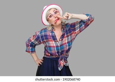 Portrait Of Funny Crazy Modern Stylish Mature Woman In Casual Style With Hat And Eyeglasses Standing With Finger Drilling Nose And Tongue Out Looking. Indoor Studio Shot Isolated On Gray Background.