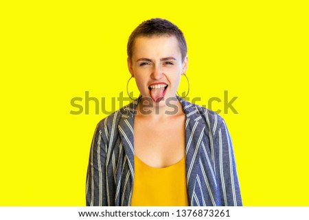Portrait of funny crazy young short hair beautiful woman in yellow shirt and striped suit standing and looking at camera with tongue out. indoor studio shot isolated on yellow background.