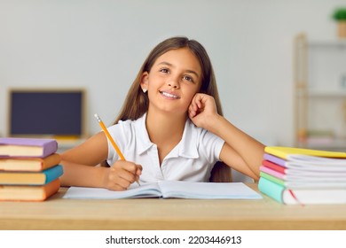Portrait of funny and cute schoolgirl with dreamy expression sitting at desk between books. Beautiful Caucasian preteen girl is thinking or dreaming while doing schoolwork in notebook.