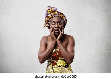 Portrait of funny emotional astonished black woman in bright clothing keeping hands on her face, astonished with gossips or big sale prices. Human facial expressions, emotions, feelings and attitude