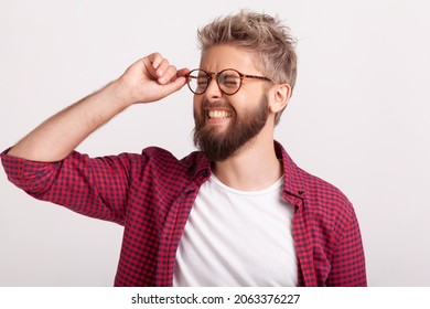 Portrait Of Funny Extremely Happy Bearded Man Touching Eyeglasses Standing With Closed Eyes And Laughing. Indoor Studio Shot Isolated On Gray Background