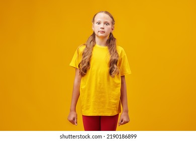 Portrait of a funny fair-haired girl child in colourful summer clothes standing motionless and looking with surprise at the camera. Studio shot on a yellow background. Children and emotions.