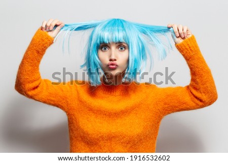 Portrait of funny girl holding to the hair of blue color, wearing  orange sweater against white background.