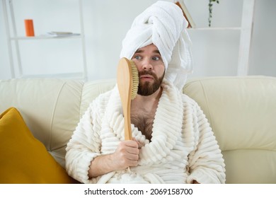 Portrait of funny guy wear turban towel holding in hands massage brush while sits on sofa. Male skin care and spa concept.