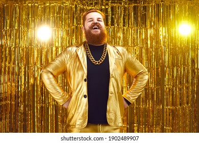 Portrait of funny happy smiling guy on stage with golden background. Content chubby bearded young man in extravagant jacket with gold chain necklace standing hands on hips and laughing