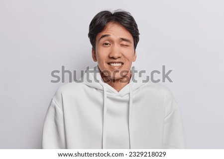 Portrait of funny Japanese man with dark hair winks eye smiles toothily dressed in casual sweatshirt feels joyful isolated over white background tries to make you laugh. People and emotions concept