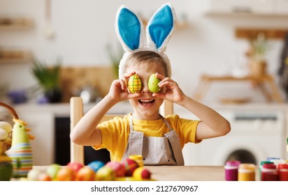 Portrait of funny kid boy with plush bunny ears on head covering eyes with multicolored Easter eggs while sitting table in cozy kitchen at home,. Easter activities for kids concept - Powered by Shutterstock