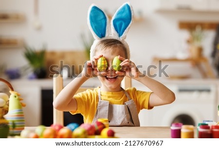 Portrait of funny kid boy with plush bunny ears on head covering eyes with multicolored Easter eggs while sitting table in cozy kitchen at home,. Easter activities for kids concept