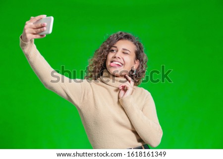 Portrait of funny kinky girl sticking out her tongue and making selfie against green background.