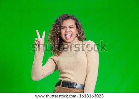 Portrait of funny kinky girl sticking out her tongue and showing peace sign at camera against green background.