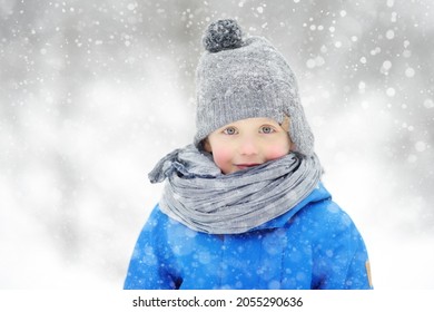 Portrait of funny little boy in blue winter clothes walks during a snowfall. Cute child wearing a warm clothing, hat, scarf is looking at you. Outdoors winter activities for family with kids.