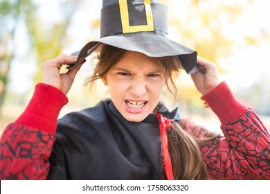 Portrait Of A Funny Little Girl Making A Sinister Face With A Paper Black Witch Hat While Walking In The Autumn Park. Halloween Meeting Concept