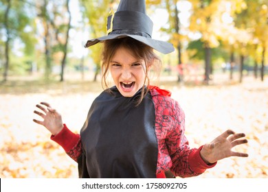 Portrait Of A Funny Little Girl Making A Sinister Face With A Paper Black Witch Hat While Walking In The Autumn Park. Halloween Meeting Concept