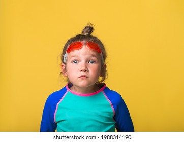 Portrait Of Funny Little Girl On Yellow Background. Kid In Swimsuit And Goggles For Swimming. Emotional Surprised Kid Face With Big Eyes Looking In Camera. Swim Or Dive In School Or Water Park.