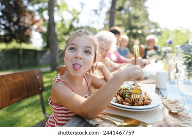 Portrait of a funny little girl sitting at table eating grilled food outdoors, sticking out tongue. Girl at family garden party. - Powered by Shutterstock