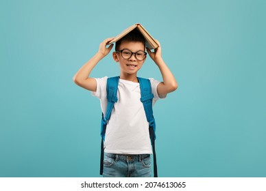 Portrait Of Funny Little Korean Schoolboy Keeping Book On Head, Cheerful Asian Male Kid Wearing Backpack Looking At Camera, Posing Over Blue Studio Background, Back To School Concept, Copy Space