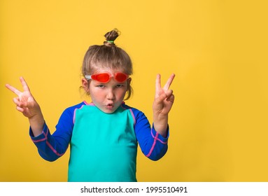 Portrait Of Funny Little Swimmer Kid Girl With Victorious Face Reaction. Victory Sign. Portrait Of Small Child In Pink Goggles And Blue Swimsuit Ready To Dive. Strong And Healthy Child On Yellow.