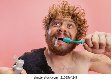 Portrait Of Funny Man In Character Of Neanderthal Brushing Teeth With Toothpaste Isolated Over Pink Background. Looks Delightful. Concept Of History, Modern And Ancient Combination, Evolution And Ad