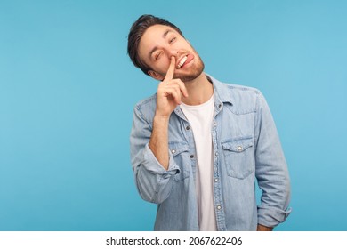 Portrait Of Funny Man Wearing Denim Shirt, Putting Finger Into His Nose And Showing Tongue, Fooling Around, Bad Habits, Disrespectful Behavior. Indoor Studio Shot Isolated On Blue Background.