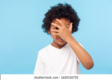 Portrait Of Funny Nosy Adorable Little Boy With Curly Hair Covering Face With Hand And Looking Through Fingers, Curious And Shy About Watching Secret. Indoor Studio Shot Isolated On Blue Background