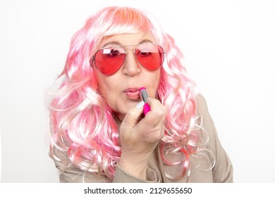 Portrait Of Funny Older Woman Painting Her Lips With Sunglasses And Pink Wig On White Background