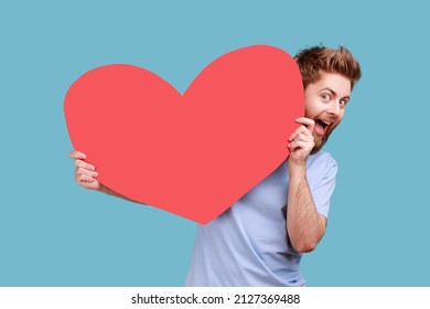 Portrait Of Funny Optimistic Bearded Man Holding Big Read Heart, Looking At Camera With Excited Facial Expression, Romantic Feelings. Indoor Studio Shot Isolated On Blue Background.