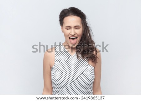 Portrait of funny positive brunette woman standing with closed eyes, showing tongue out, demonstrates childish behavior, wearing striped dress. Indoor studio shot isolated on gray background.