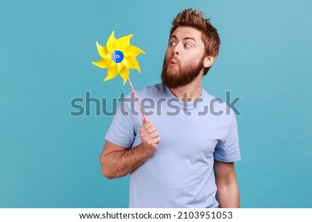 Portrait of funny positive childish handsome bearded man wearing T-shirt playing blowing yellow windmill, having fun with paper toy. Indoor studio shot isolated on blue background.
