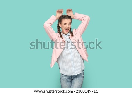 Portrait of funny positive childish teenager girl with braids wearing pink jacket showing bunny ears, winking to camera. Indoor studio shot isolated on green background.