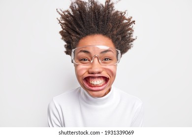 Portrait of funny positive dark skinned woman smiles with teeth has big mouth poses under air pressure has hair standing up dressed in big transparent glasses isolated on white wall