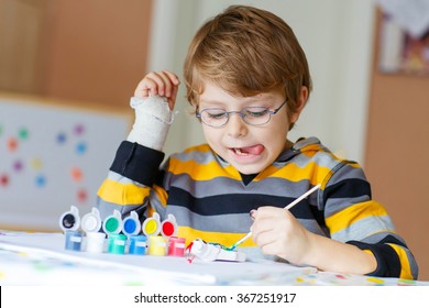 Portrait Of Funny Preschool Kid Boy With Glasses And Broken Hand Drawing. Little Child Having Fun With Colorful Watercolors And Gouache, Indoors. School, Education Concept