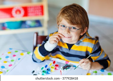 Portrait Of Funny Preschool Kid Boy With Glasses And Broken Hand Drawing. Little Child Having Fun With Colorful Watercolors And Gouache, Indoors. School, Education Concept